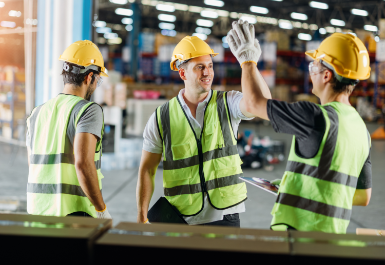 Workers in hi-vis vests, safety gloves and hard hats high-five on the factory floor
